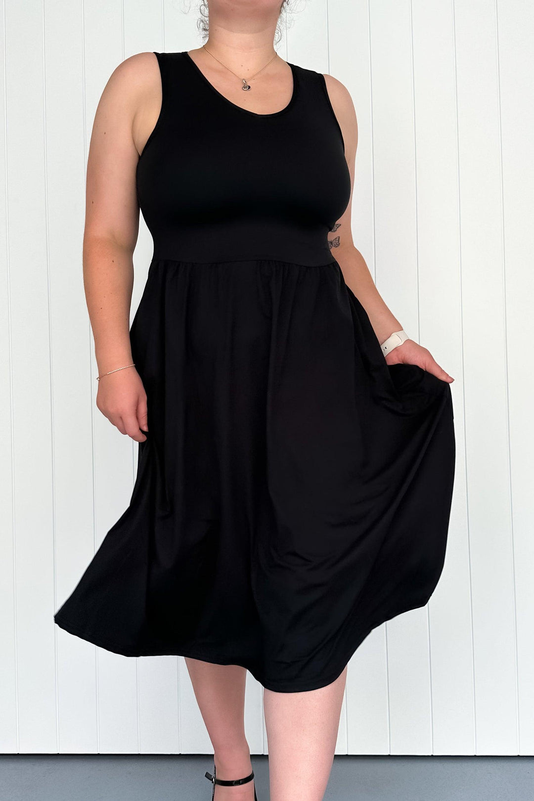 A model wearing a sleeveless black midi dress with pockets, standing against a white background.
