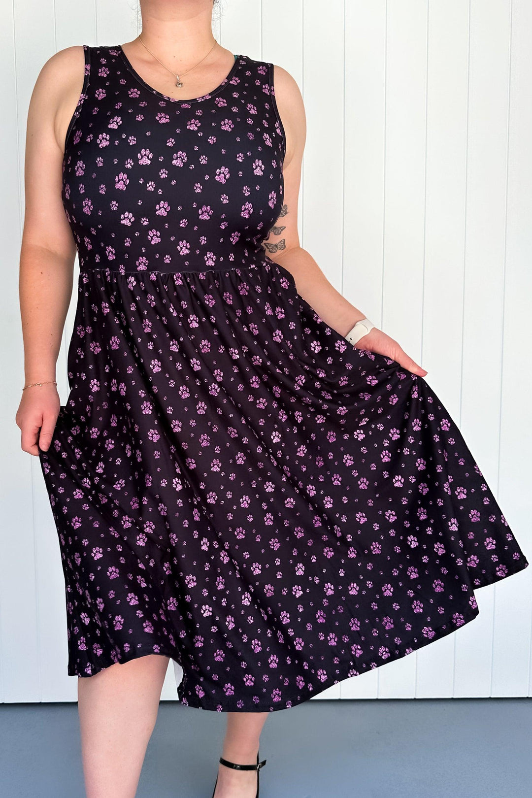 A sleeveless midi dress with a deep black base scattered with tiny paw prints in soft pinks and purples. The dress has a round neckline and deep side pockets.