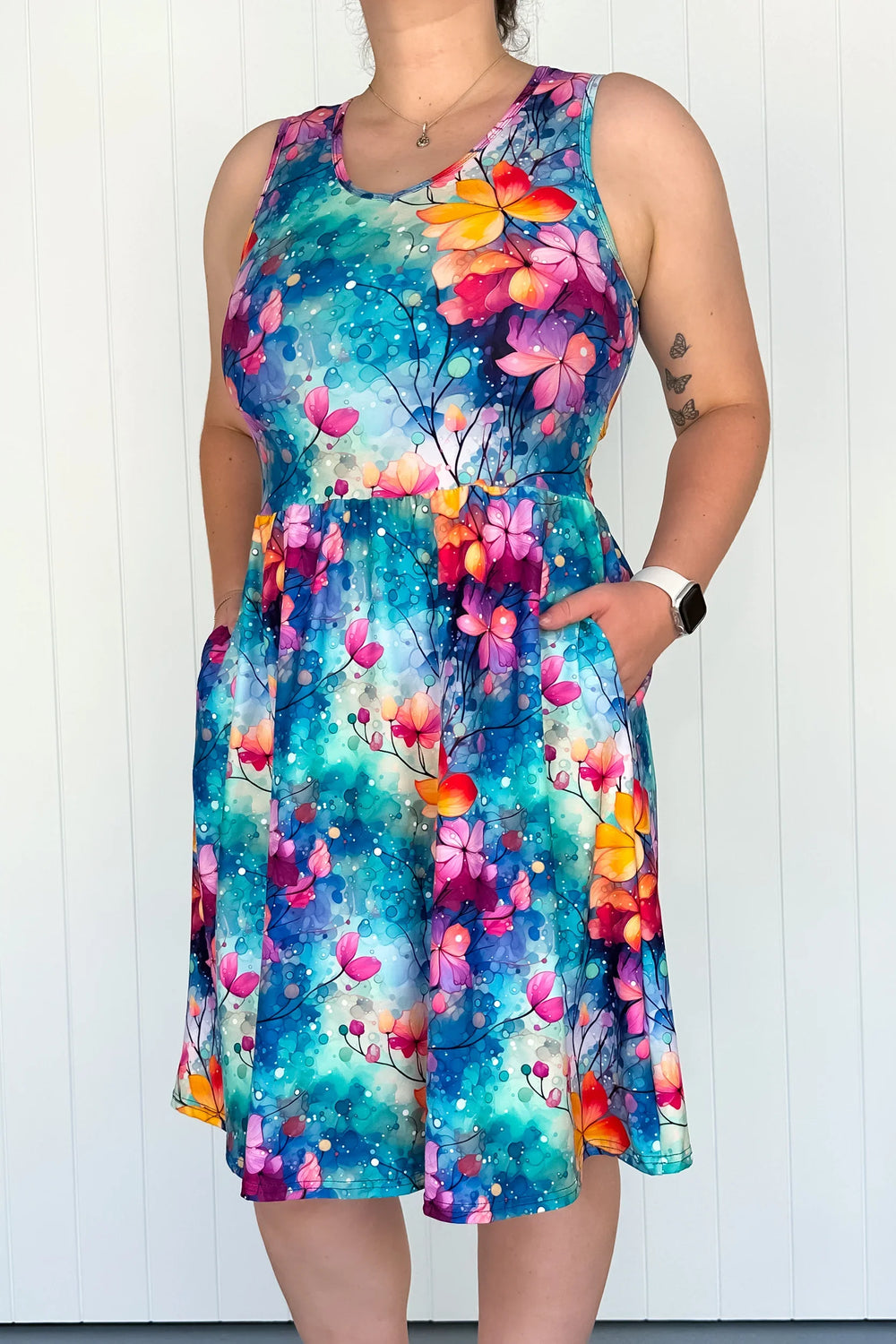 A sleeveless knee-length floral dress with side pockets, ocean-blue background with vibrant colors of pink, orange, and green from the floral pattern.