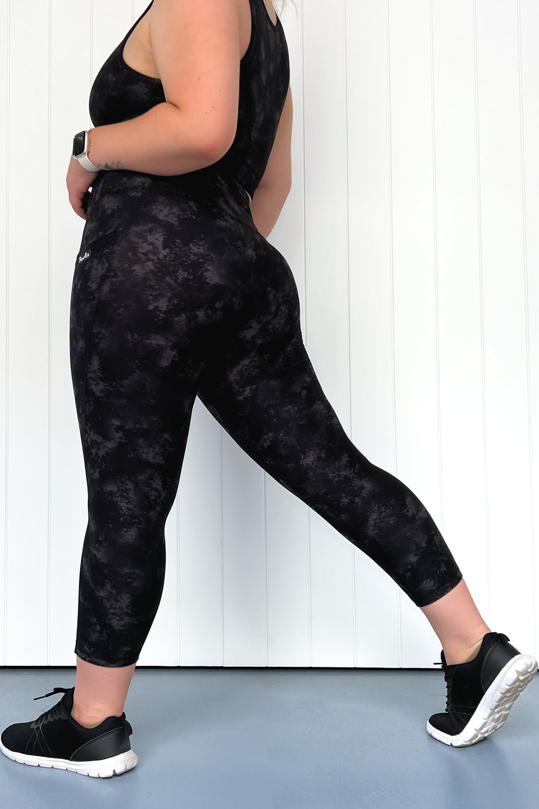Person wearing black tie-dye leggings and black sneakers on a light blue floor with white wall background