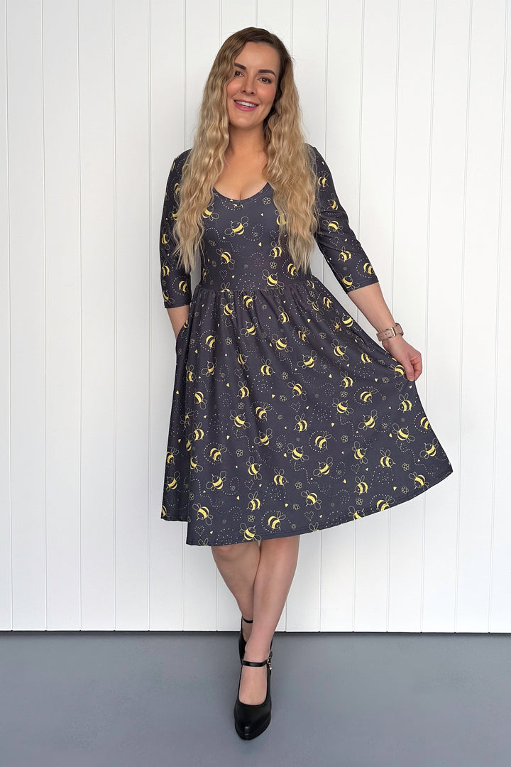 Woman wearing a dark grey dress with yellow bee pattern against a white wooden panel background